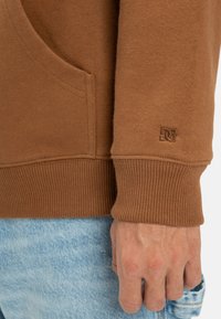 Brown sweatshirt with ribbed cuffs, featuring a side pocket and a small embossed logo. Worn over light blue jeans.