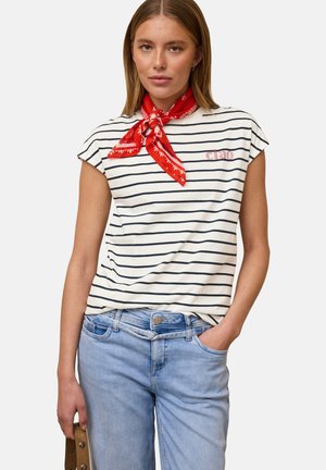 Kvinde iført en hvid og sort stribet skjorte med teksten "ciao", rødt halstørklæde, lyseblå jeans, der holder en brun pung i venstre hånd.