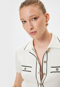 Polo shirt bianco a coste con rifiniture nere a contrasto, maniche corte, due taschini sul petto e bottoni dorati. Ingrandimento della zona del colletto.