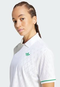 Camisa polo blanca con patrón de diamantes, logo verde de tres rayas en el lado izquierdo del pecho y acentos verdes en el cuello y las mangas.