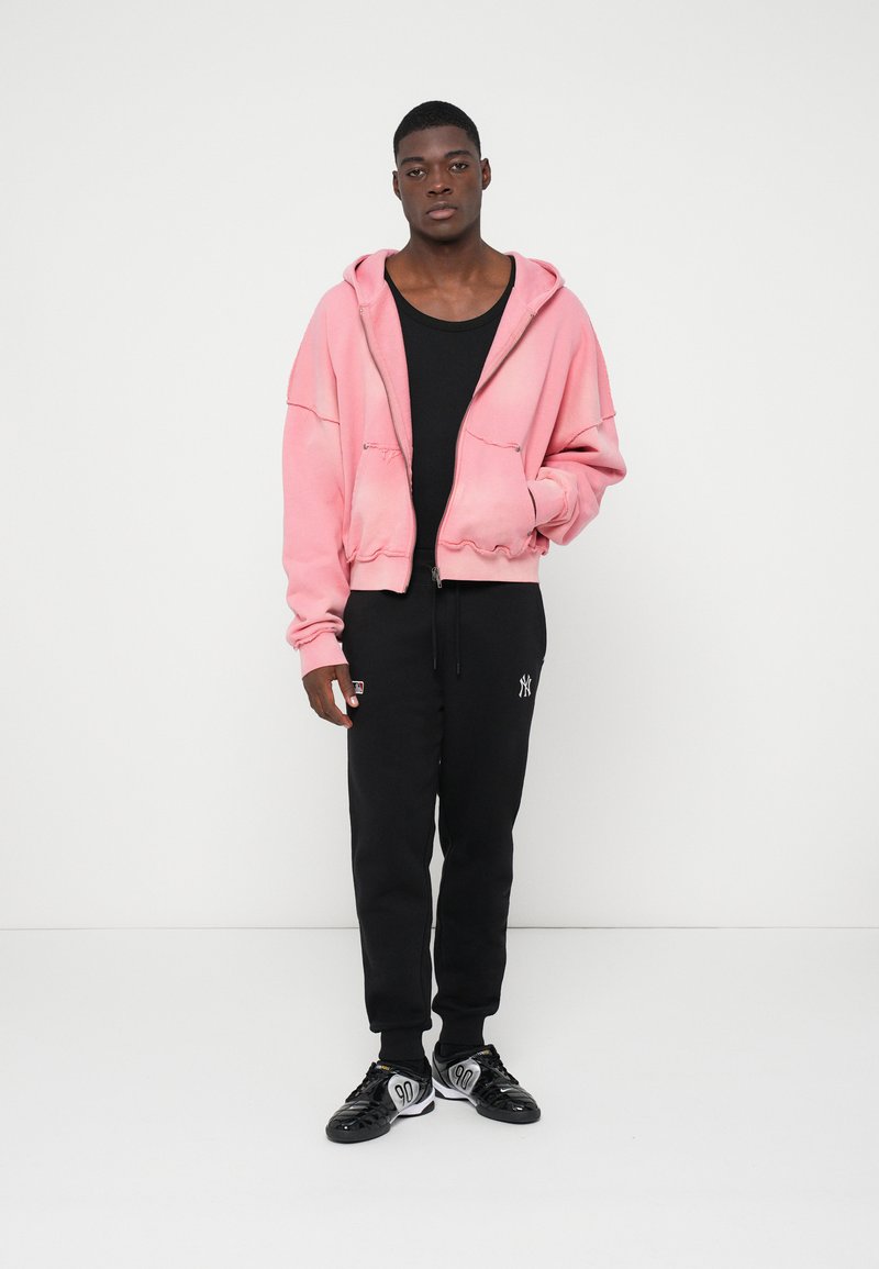Pink oversized zip-up hoodie with a front pocket, worn over a black fitted shirt, paired with black sweatpants featuring a logo. Black sneakers.