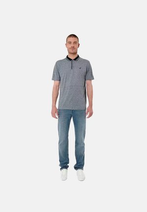 Man staat met het gezicht naar voren, draagt een grijs poloshirt, blauwe spijkerbroek en witte sneakers tegen een effen lichte achtergrond.