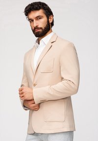 Homme aux cheveux bouclés foncés et à la barbe, portant un blazer beige sur une chemise blanche, ajustant sa manche devant un fond clair uni.