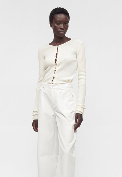 Γυναίκα φοράει κρεμ ριμπ πλεκτή ζακέτα με κουμπιά και λευκό παντελόνι με ψηλή μέση και φαρδύ πόδι, στέκεται μπροστά σε απλό λευκό φόντο.