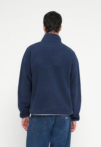 Granatowy polarowy pulower z wysokim kołnierzem, o fakturowanym wykończeniu i luźnym kroju. Posiada szew na górnej części pleców oraz elastyczne mankiety.