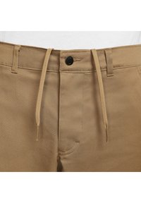 Närbild av beige byxor vid midjan med bältesöglor, svart knapp och beige snörning hängande ner mot en vit bakgrund.