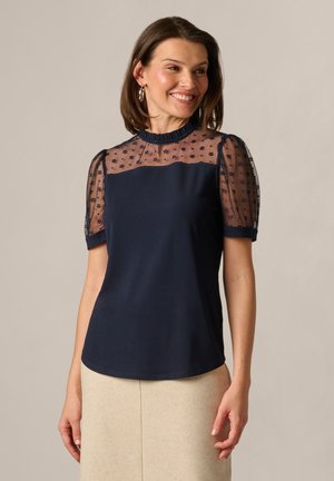 Frau trägt eine marineblaue Bluse mit transparentem, floraler Stickerei am Einsatz und Puffärmeln, kombiniert mit einem beigen Rock, lächelt und schaut nach rechts.