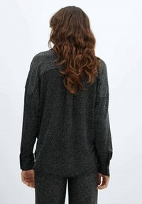 Personne avec de longs cheveux bruns ondulés, portant un haut à manches longues gris foncé avec des speckles et un pantalon assorti, de dos sur un fond clair.