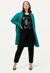 Teal lång kappa över en svart paljetterad grafisk t-shirt, i kombination med svarta byxor och silvriga högklackade skor. Svala texturer genom hela looken.