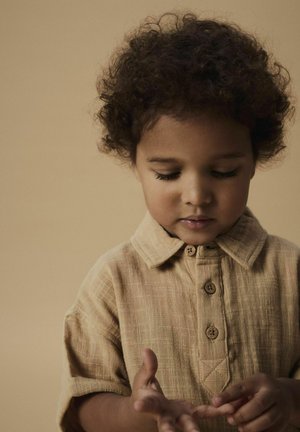 Beige shirt met korte mouwen, voorzien van een knopenpad, kraag en een textuurstof. Kind houdt handen samen, kijkend naar beneden. Zacht, golvend haar.