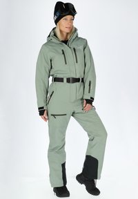 Light green ski suit with black accents, featuring zippered pockets, a belt, and reinforced cuffs. Worn with a black beanie and ski goggles.