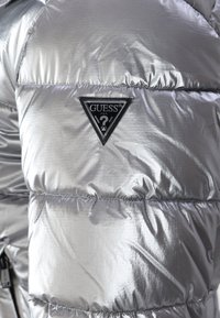 Giubbotto imbottito argento con una superficie lucida e testurizzata. Presenta un patch logo triangolare nero con il nome del marchio "GUESS."