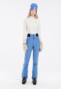 Lichtblauwe skibroek met hoge taille, zwarte accenten en drie knopen. Een wit lang mouwtopje en beige wanten maken de outfit compleet.