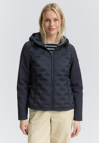Donna che indossa un giubbotto trapuntato nero con cappuccio e zip, una maglietta a righe sotto, e pantaloni beige chiaro, in piedi contro uno sfondo uniforme.
