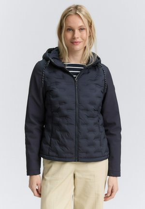Femme portant une veste noire matelassée avec capuche et zip, un chemisier rayé en dessous, et un pantalon beige clair, se tenant devant un fond uni.