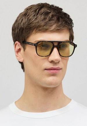 Junger Mann mit kurzen braunen Haaren, der rechteckige Schildpatt-Sonnenbrillen mit gelb getönten Gläsern und ein weißes Rundhalsshirt trägt.