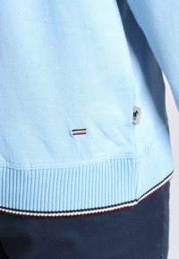 Pull léger bleu clair avec un ourlet côtelé. Présente une petite étiquette de logo et un accent rayé en marine, rouge et blanc au niveau du poignet. Matière en mélange de coton.