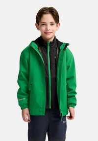 Groene waterdichte jas met een capuchon, voorzien van een zwart ritsvest aan de binnenkant, reflecterende accenten en elastische manchetten. Draagt over donkere broek.