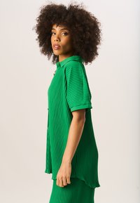 Green pleated shirt featuring short sleeves, a pointed collar, and button closure at the front. The fabric has a textured, ribbed design.