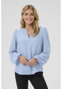 Blouse bleu clair avec un décolleté en V et des manches bouffantes. Fabriquée en tissu texturé, elle présente une coupe décontractée et des poignets élastiques. Pantalons noirs visibles.