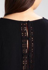 Haut noir en tissu texturé, doté d'une accent en dentelle le long de l'ouverture du dos, fermé par un bouton au cou.