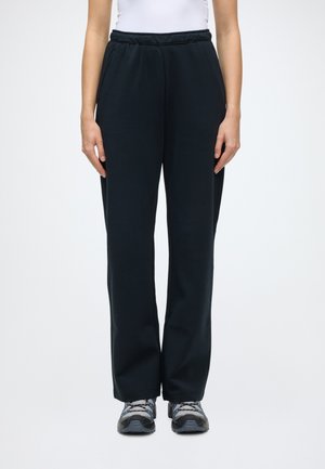 Personne portant un pantalon noir droit, un haut blanc et des chaussures de randonnée grises, debout devant un fond clair uni.