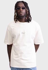Cremfarbener Baumwoll-T-Shirt mit kurzen Ärmeln, rundem Halsausschnitt und gesticktem Text "Rebel" in Kursivschrift auf der Brust.