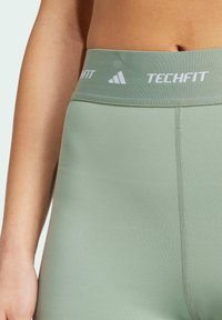 Green athletic leggings with a wide elastic waistband labeled "TECHFIT." Features smooth texture and flat seams for comfort.