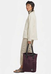 Maroon tote bag with black straps, made of durable fabric. Features a front pocket and reinforced stitching. Person wears a cream fleece jacket.