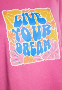 T-shirt rosa con una grafica centrale raffigurante la scritta "VIVI IL TUO SOGNO" in blu, circondata da motivi floreali colorati e accenti.
