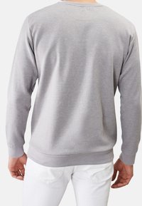 Uomo che indossa una semplice maglietta grigia chiara a maniche lunghe e pantaloni bianchi, visto da dietro su uno sfondo bianco.