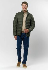 Zelená prešívaná bunda s stojatým golierom, nosená cez béžovú košeľu. Model stojí v džínsoch a hnedých teniskách na neutrálnom pozadí.