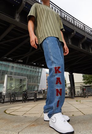 Person wearing striped olive green shirt, blue jeans with red "KANI" letters, and white Nike sneakers, standing under a metal bridge in urban setting.