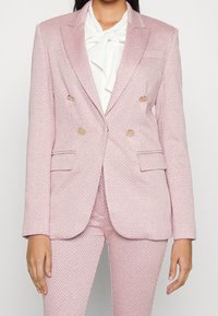 Blazer doppiopetto rosa chiaro con motivo a spina di pesce, bottoni dorati, colletto a risvolto e due tasche frontali. Abbinato a una camicia bianca.