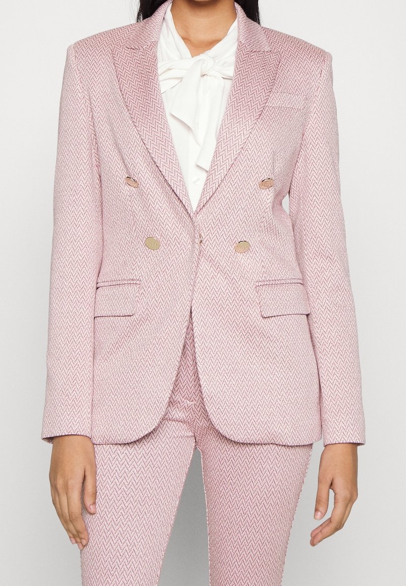Blazer doppiopetto rosa chiaro con motivo a spina di pesce, bottoni dorati, colletto a risvolto e due tasche frontali. Abbinato a una camicia bianca.