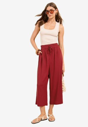 Frau trägt beige ärmelloses Oberteil, hoch taillierte, lockere rote Hose, beigen Sandalen, Sonnenbrille und hält eine gewebte Handtasche, steht vor weißem Hintergrund.