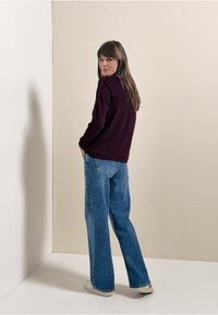 Bordeaux lange mouw top gecombineerd met lichtblauwe wijde jeans, staand tegen een neutrale achtergrond. Ivoorkleurige sneakers maken de outfit compleet.