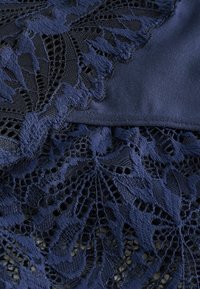 Țesătură din dantelă bleumarin cu modele complexe de frunze, suprapusă peste o țesătură netedă, mai închisă la culoare. Texturile se contrastază proeminent.