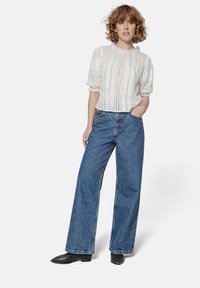 White textured blouse with ruffled neckline, paired with high-waisted, loose-fitting blue denim jeans. Model wears black ankle boots.
