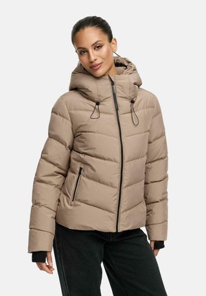 Kvinde iført en beige vatteret jakke med hætte og sorte lynlåslommer, kombineret med sorte bukser mod en ensfarvet baggrund.