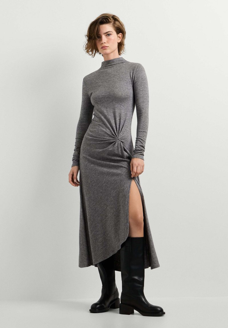 Femme portant une robe grise à manches longues avec une fente latérale et un détail froncé, associée à des bottes noires montantes jusqu’aux genoux, debout devant un fond uni.
