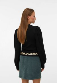 Pull en noir texturé avec des manches bouffantes et une taille rayée beige, associé à une jupe en velours côtelé verte.