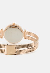 Roza zlata mrežasta ura z okroglo škatlo in prozornim steklom. Ima hinge zapiralo in gladek kovinski zaključek. Zadaj so prikazane podrobnosti blagovne znamke.