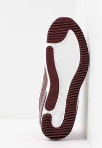 Sula av maroon och vit sportskor med Nike Air Max-logotyp, som visar mönstret på sulan och Nike-swoosh, mot vita hörnväggar.