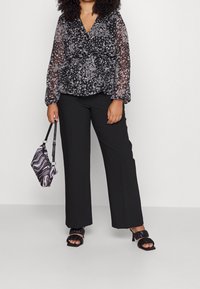Krøllet hårmodel står selvsikkert i en blomsterpræget, semigennemsigtig bluse, brede sorte bukser og stilfulde sorte hæle, mens hun holder en mønstrede taske.