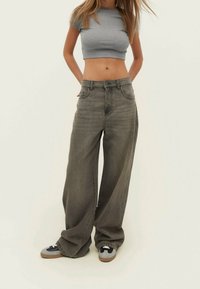 Mujer con un top corto gris, jeans gris sueltos y zapatillas, de pie con las manos detrás de la espalda contra un fondo claro y liso.