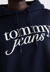 Felpa blu navy realizzata in morbido tessuto, con un grande logo bianco "tommy jeans" sulla parte anteriore. Include coulisse regolabili.