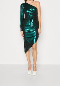 Metallisk smaragdgrön klänning med en lång ärm, sidoknytte och asymmetrisk fåll. Matchas med vita platåskor. Slät textur.