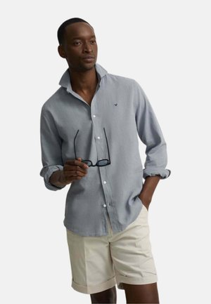 Hombre con camisa azul clara de botones y mangas remangadas, pantalones cortos beige, sosteniendo gafas negras, de pie con una mano en el bolsillo.