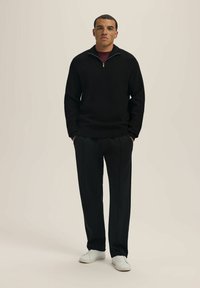 Czarny sweter dzianinowy z zamkiem przy kołnierzu, w połączeniu z czarnymi spódnicami plisowanymi i białymi sneakersami. Model stoi z rękami w kieszeniach na neutralnym tle.